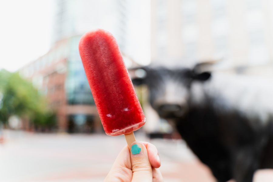 A hand holds up a red frozen treat from Locopops with the bull statue in Downtown Durham in the background.