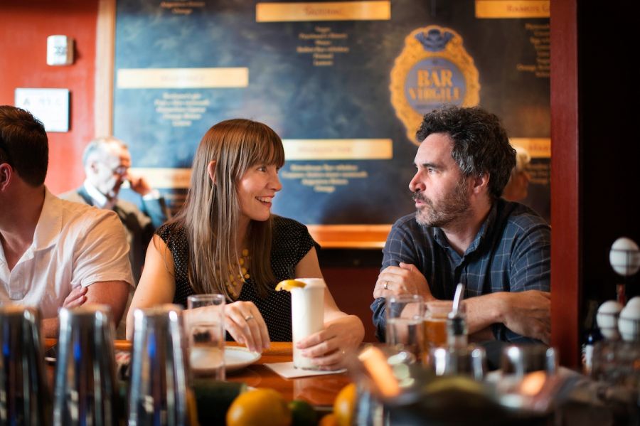 A man and woman in conversation while sitting at a bar. The woman has a white drink in a tall glass. They are smiling.
