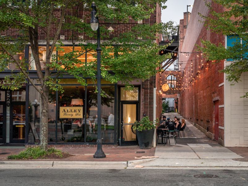 A photo of a restaurant in downtown from across the street. People dining inside and outside in the alley next door.