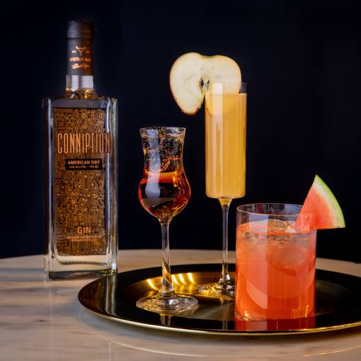 A bottle of conniption Gin stands next to two colorful cocktails on a table.