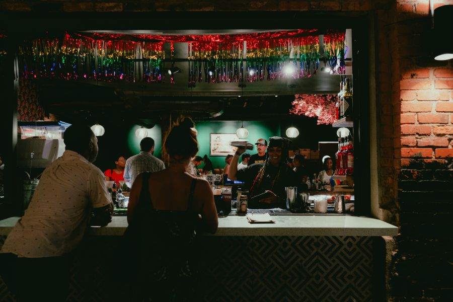 Two adults lean on the counter on the outdoor patio while a bartender shakes a cocktail and smiles.
