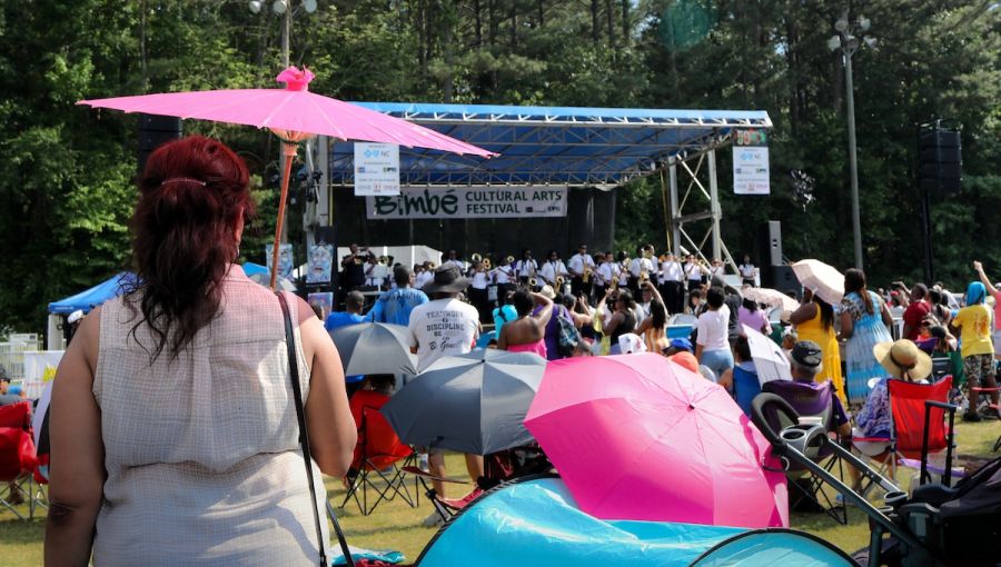 Concert goers at Bimbé Cultural Arts Festival hold pink umbrellas over their heads in the sun as they enjoy live music.