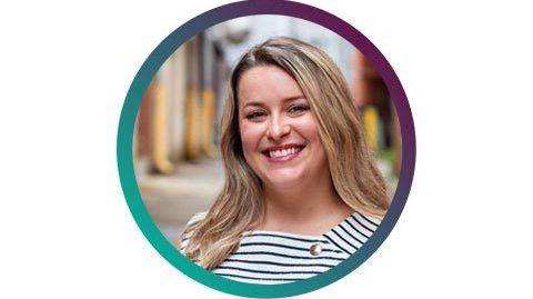 A blonde woman wearing a blue and white striped shirt smiles at the camera.