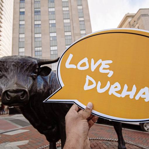 A speech bubble that says "Love, Durham" being held in front of the Major the Bull statue. City streets in the background.