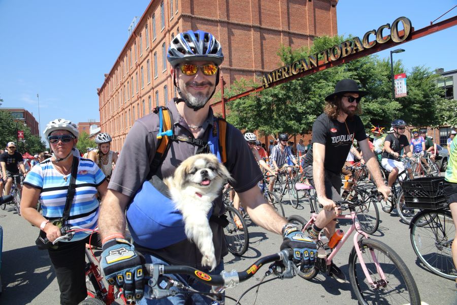 A group of people on bicycles. Foreground: a man wearing a helmet and sunglasses smiles at the camera with a dog in a bag.