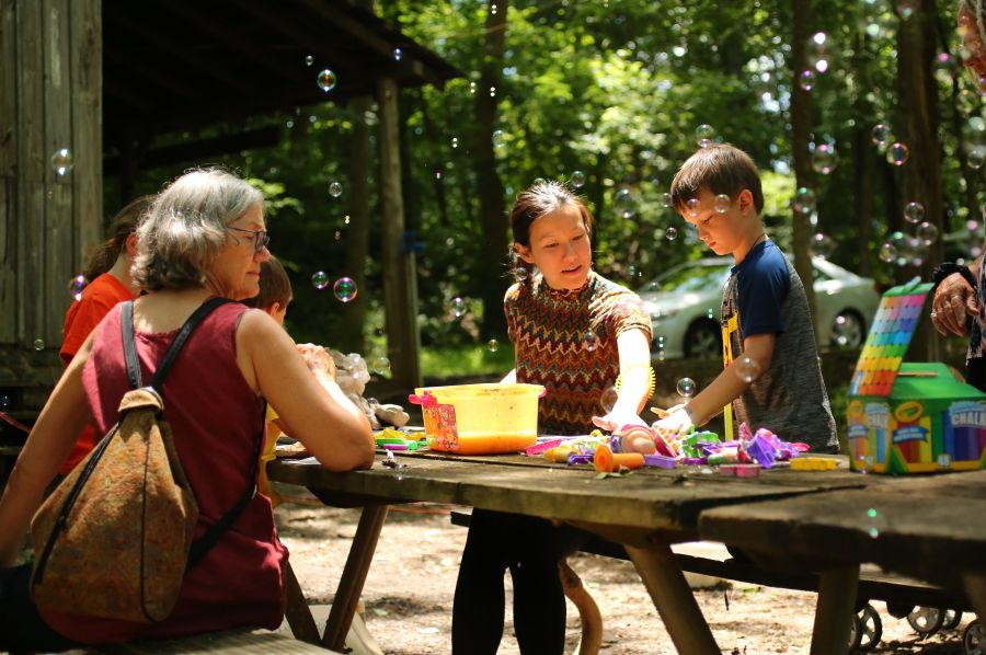 A mother and her son enjoy crafts at an outdoor picnic table while bubbles float through the air.