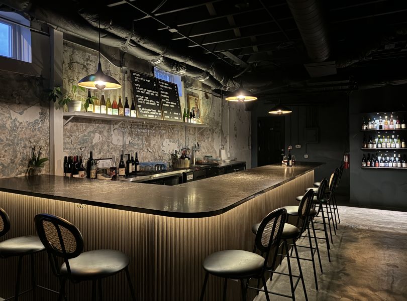 A dimly lit bar with a black top and black bar stools, wine bottles and glasses line the back bar.