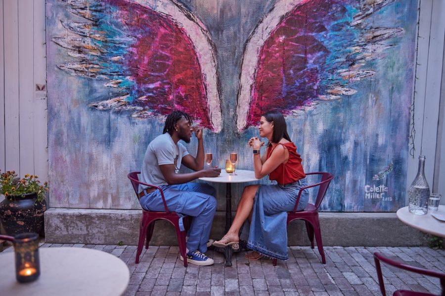 A couple shares a bottle of wine at an outdoor table in front of a pink, blue and purple mural of wings.