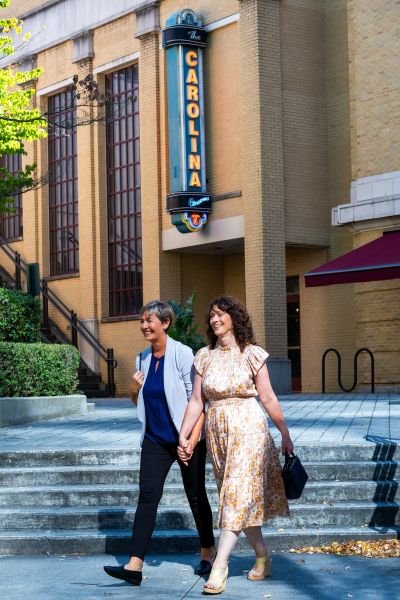 Two women walk hand-in-hand outside. Behind them, there's a building with a lit sign for the Carolina Theatre.