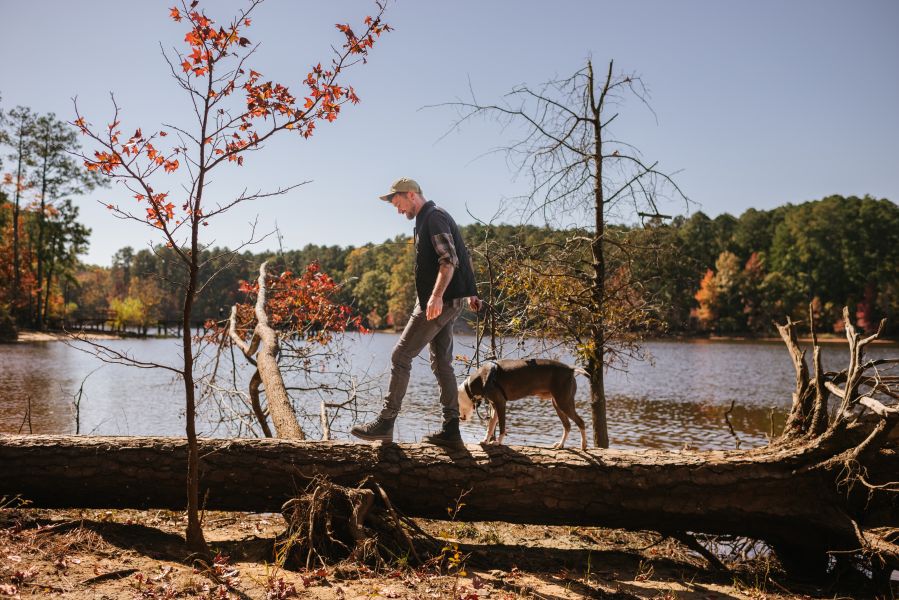 A man and a dog balance on a fallen tree with Falls Lake in the background.