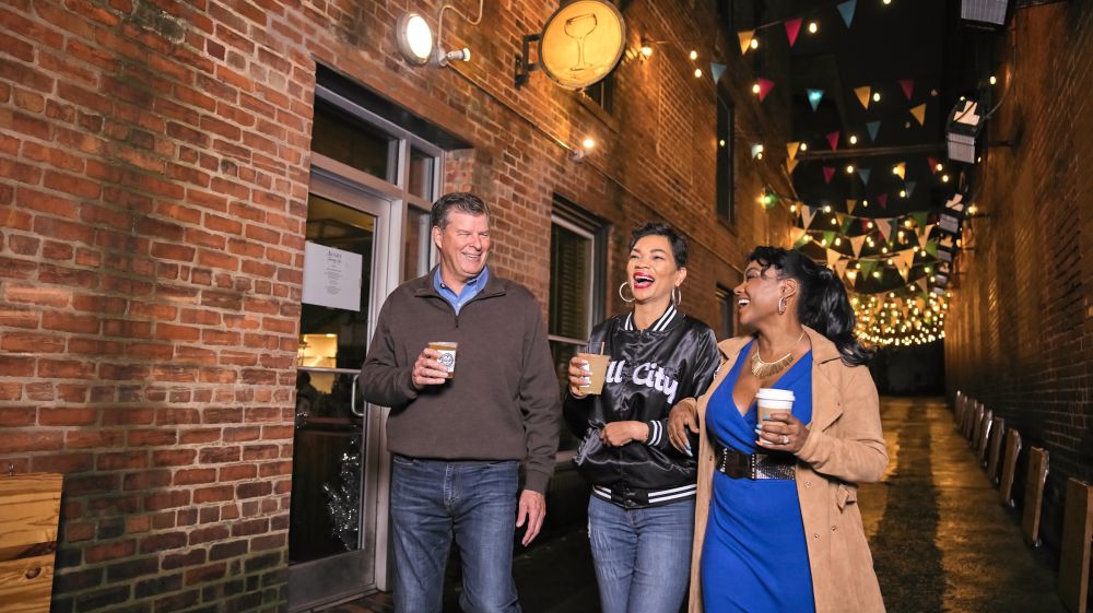 Three friends walking under twinkle lights and laughing with to-go drinks.