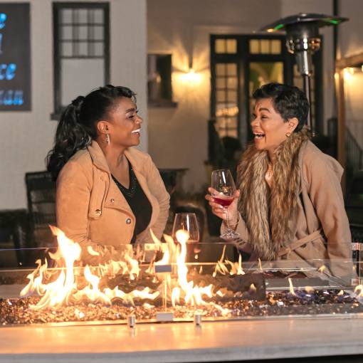 Two women laugh and share drinks by an indoor fire pit.