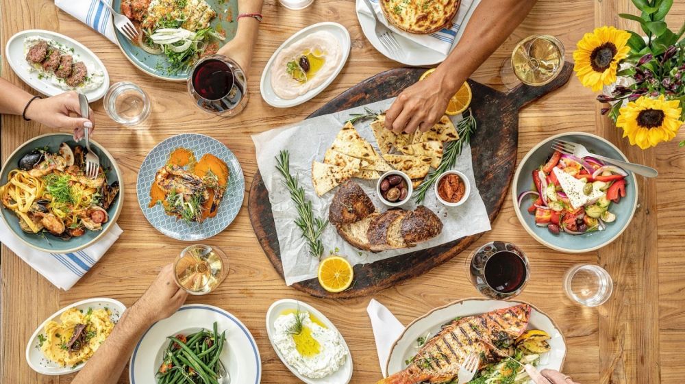 a table full of colorful dishes of food and people reaching for glasses and portions of food, shot from above