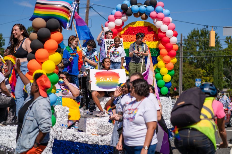 A young person surrounded by fellow parade participants sits on the back of a parade float, holding a sign that says “Slay.”