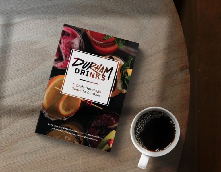 A booklet called Durham Drinks: A Craft Beverage Guide to Durham sits on a table next to a cup of coffee.